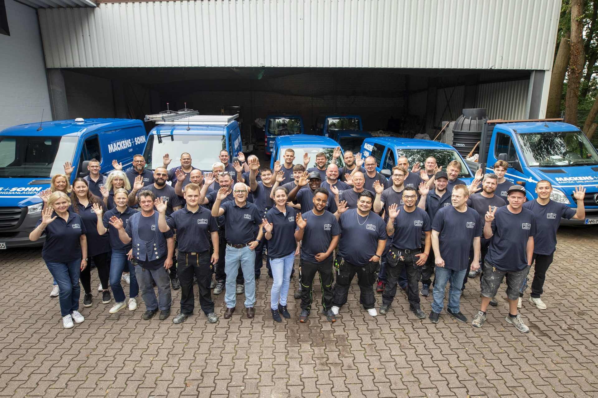 Das Team von Mackens + Sohn winkt vor der blauen Fahrzeugflotte